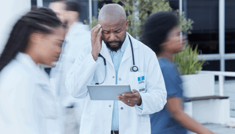 A doctor holding his head while looking at a tablet as other healthcare professionals walk past.